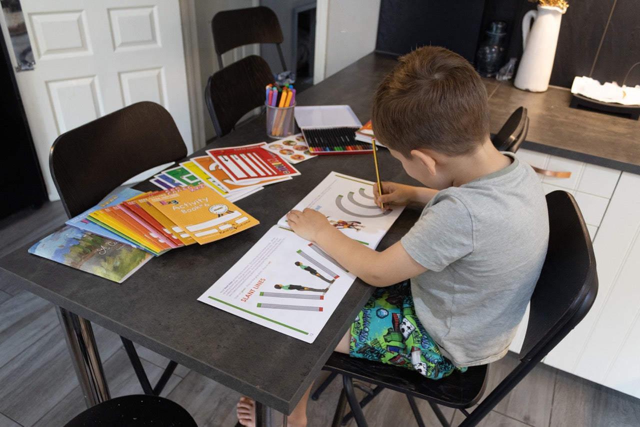 Child seated at a table completing pre-writing stroke activities with a pencil while using handwriting workbooks and learning materials.