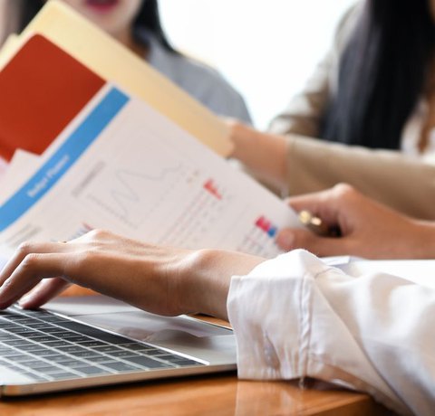 Three people collaborate at a table, focused on the February 25th & 26th, 2025 LIVE SMHP and TIER ONE Therapist Certification Webinars by Real OT Solutions. One types on a laptop, another holds a folder, and the third points at a printed graph, highlighting their hands and documents.