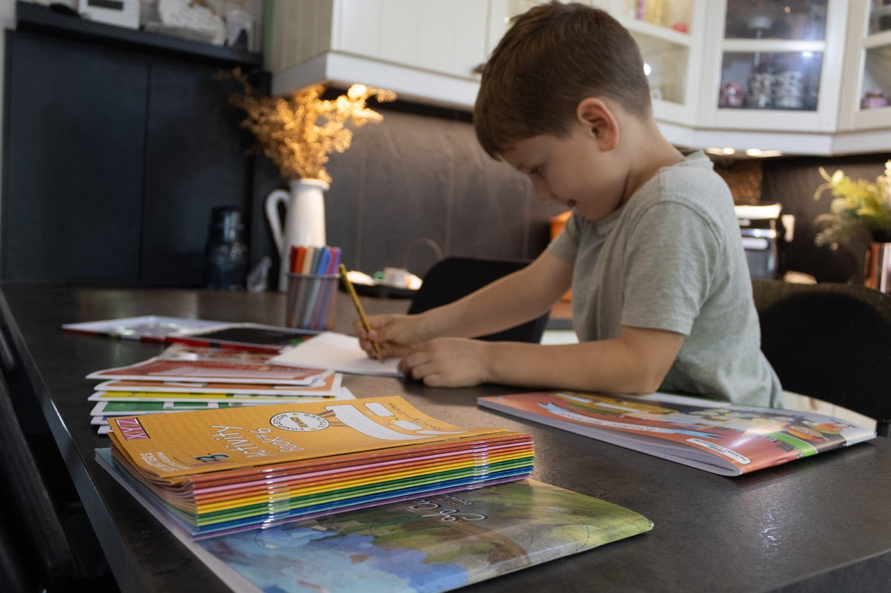 Young child focused on handwriting practice at a table with activity books, workbooks, and writing materials nearby.