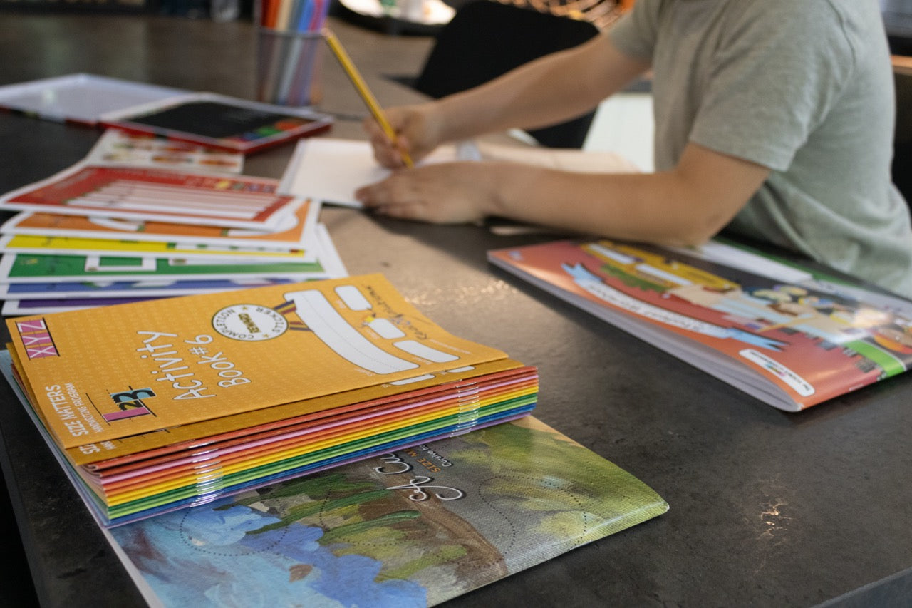 Child practicing handwriting with a pencil at a table alongside colorful activity books and handwriting learning materials.